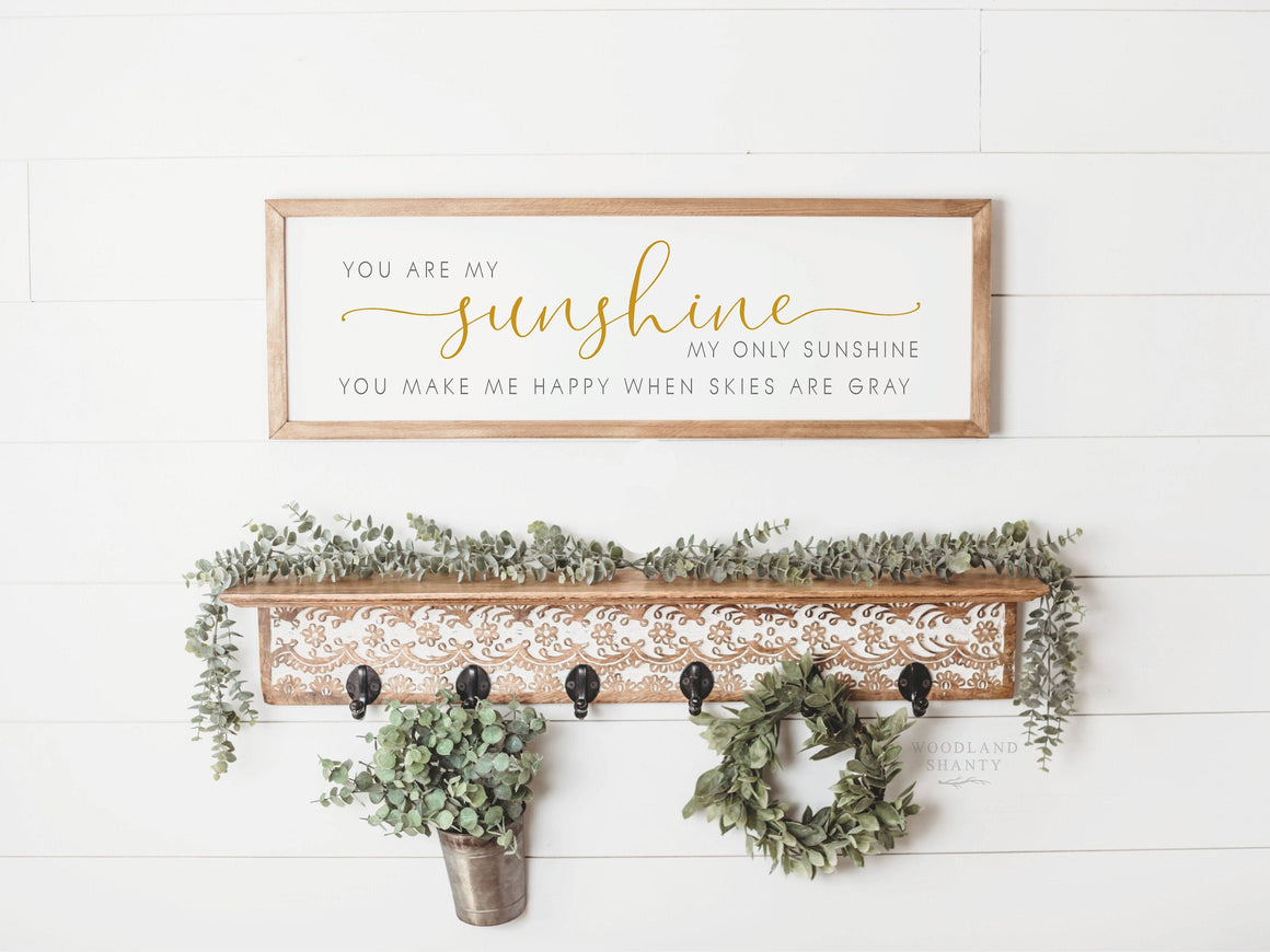 May include: A wooden sign with the text "You are my sunshine, my only sunshine, you make me happy when skies are gray". The sign is hanging on a white wall with a shelf underneath. The shelf has a decorative design and is decorated with greenery.