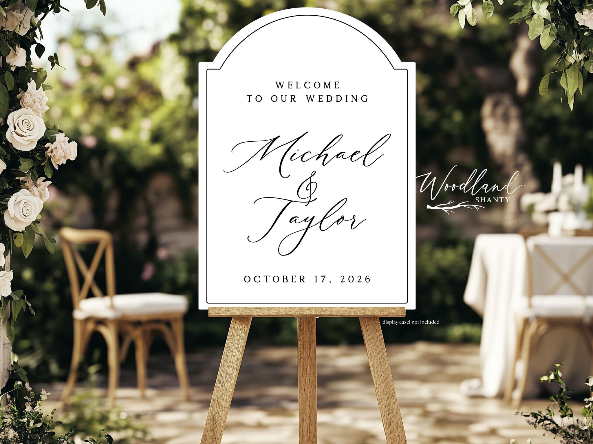 May include: A white wedding welcome sign on a wooden easel. The sign has a decorative arched top and features the names "Michael & Taylor" and the date "October 17, 2026" in elegant script. The background is a blurred outdoor setting.