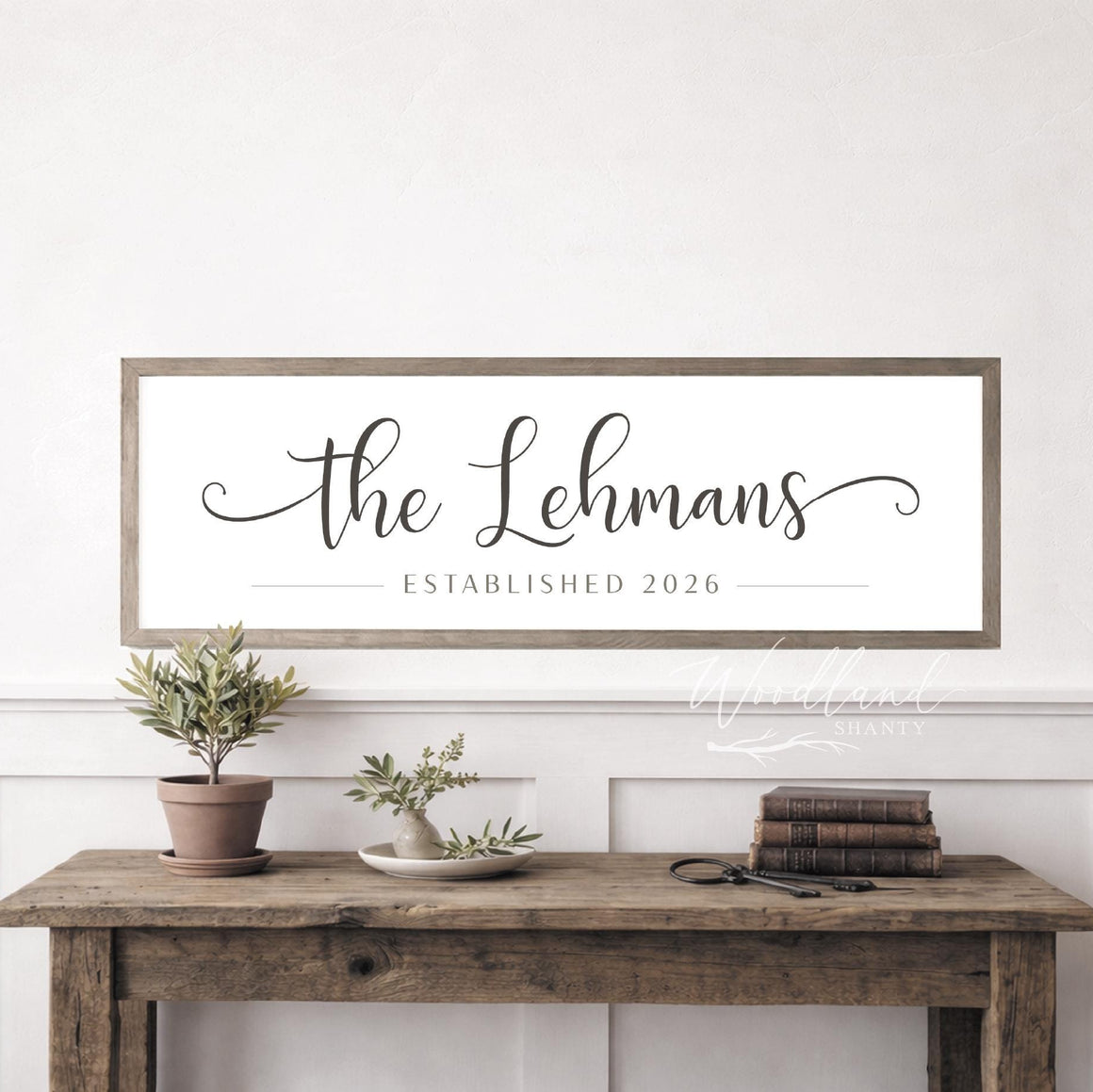 May include: A rectangular wooden sign with the text "the Lehmans" in elegant script, with "ESTABLISHED 2026" below. The sign is framed and hangs above a rustic wooden table with plants and books.