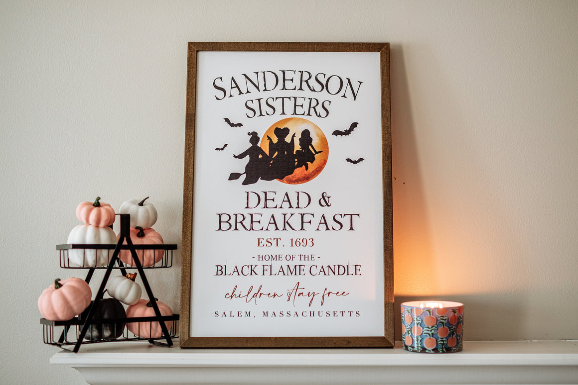 May include: A framed sign with the text "Sanderson Sisters Dead & Breakfast Est. 1693 - Home of the Black Flame Candle children stay free Salem, Massachusetts". The sign features a black silhouette of three witches flying on a broomstick in front of a full moon. The sign is on a white mantel with a candle and a tiered stand with pink and white pumpkins.